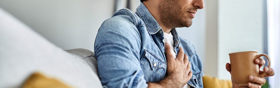 Young man feeling sick and holding his chest in pain while drinking tea in the living room.